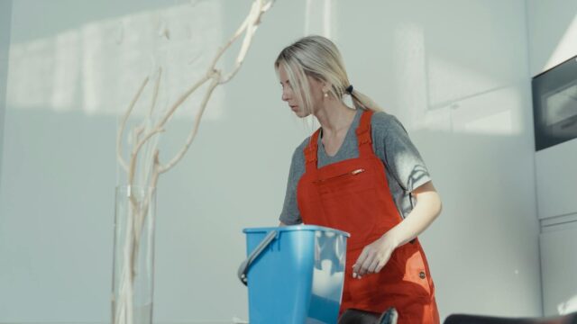 A woman in a red uniform cleans indoors with a blue bucket, ensuring a spotless environment.