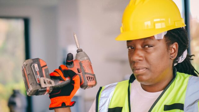 Black female construction worker with a drill, wearing safety equipment and hard hat.